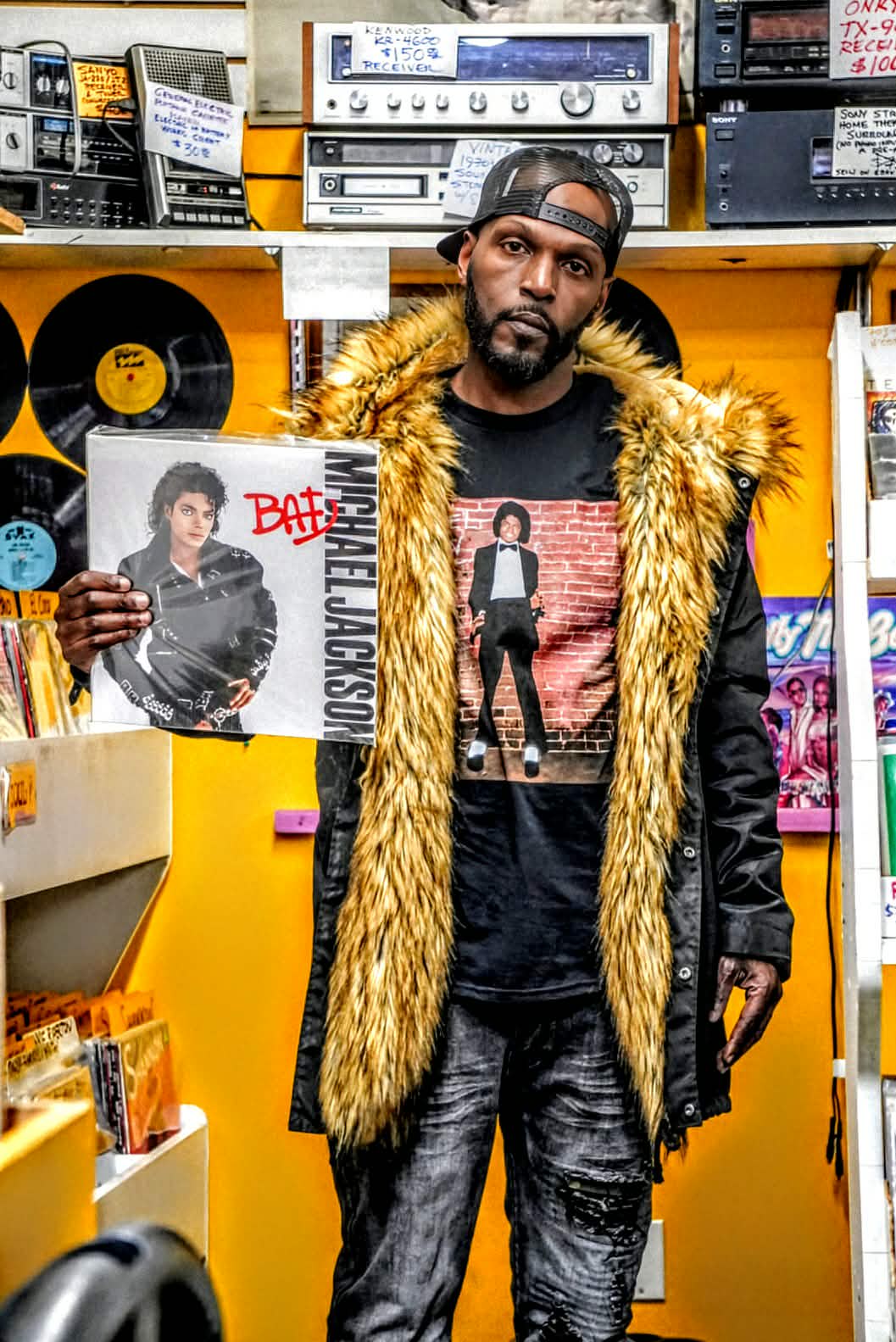 Wiz Hoffa facing the camera in a record store, holding Michael Jackson's 'Bad' vinyl, gold fur-collar parka over an MJ tee, with a wall of records and vintage receivers behind him
