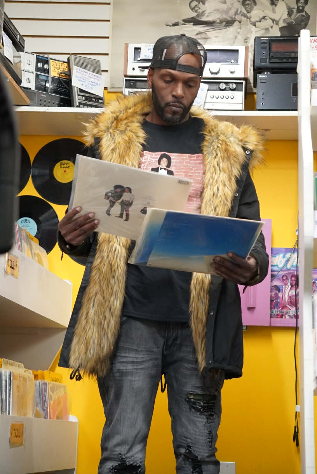 Wiz Hoffa standing in a record store, examining two vinyl LPs, one in each hand, fur-collar parka on, wall of vinyl behind him
